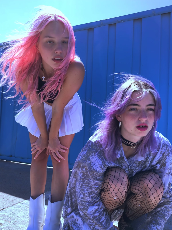 A candid snapshot of two young women with vibrantly colored pastel hair—one with soft pink loose waves, the other with lavender tousled layers—posed in front of a blue construction wall on a sunny day. The first leans forward wearing a crisp white tennis skirt and chunky platform boots; the second crouches in a loose printed shirt, black fishnets, and subtle chokers. Both have minimal, natural makeup with lips slightly parted, wearing blank yet camera-ready expressions. Muted daylight hits the scene with slight sun flare reminiscent of a subtle digital glitch, casting cool-toned bluish-purple shadows. Textural details highlight visible skin imperfections, intricately detailed fabric wrinkles and fishnet patterns, and individually defined pastel hair strands. The framing is casually off-center and slightly tilted, capturing the spontaneous yet intimate attitude of mid-2010s Tumblr-style grunge photography, intentionally softened with muted saturation and lowered contrast throughout.