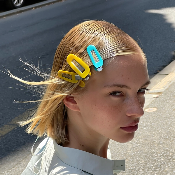 Bright yellow and cyan thick plastic clips line one side of sleek, blonde hair slicked back behind the ears, catching the crisp natural daylight that gently highlights the subtle pores and faint freckles on her skin. The clips shine with soft reflections, framing a calm stare that meets the camera just as a slight breeze teases a few stray strands free. A lightweight shirt collar peeks into the frame with loose threads barely visible, while the photo tilts slightly from a high arm stretch, grounding the scene near a sunlit sidewalk dotted with city stones—authentic iPhone close-up, statement hair-clip focus