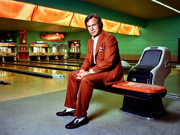 In the smoky, neon-glow ambiance of a 1970s bowling alley, a white man sits poised on a sleek, vinyl-tufted retro barstool. His rust-colored three-piece suit—fitted with a snug waistcoat and pressed trousers—reflects the warm amber hues of the sodium-vapour lamps hanging low overhead, casting elongated shadows across the polished, worn wood lanes. His wavy chestnut hair, parted neatly but with natural body, frames thick sideburns that brush the collar of his crisp, ivory shirt. The subtle sheen of his soft leather loafers catches the faint, flickering reflections from coloured pinspotters, while a confident, unflinching stare challenges the viewer’s gaze, full of quiet authority and cool self-assurance.

The camera’s eye-level 50 mm lens captures this moment with intimate clarity, soft grain echoing the analog aesthetic of Kodak 5247 film stock. The film grain whispers against the rich textures of the man’s tailored fabric, the smooth gloss of barstool vinyl, and the marbled surface of the scoring counters behind him. His skin pores and subtle stubble come alive under the meticulous directional lighting, grounding the image in tactile realism. The background blurs delicately, the lurid greens and deep siennas of the bowling alley décor dissolving into warm, nostalgic hues.

The composition balances the subject slightly off-centre, inviting the viewer to both focus on his magnetic expression and absorb the environment's retro charm—from the vintage pinsetters to the tiled floor with its cracked grout. Everything pulses with the subdued energy of a forgotten era, textured with the visual language of classic late 70s cinema, capturing a moment that feels both real and cinematic. This is a snapshot of cool confidence embedded in the golden haze of time.

—late-70s / early-80s cinematic photograph, authentic film grain.