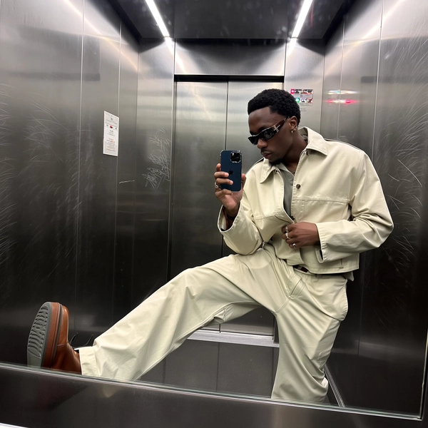 Cool LED strips hum overhead, casting a gentle sheen across the brushed steel walls etched with whispered graffiti scars, as he settles low on the elevator floor, knee drawn up with an ease that feels grounded yet decidedly luxurious. His light utility co-ord falls in fluid, tailored folds of soft cotton-blend gabardine, the sculptural sunglasses perched with intentional edge catching muted reflections. The phone rests just below chest height, angled slightly askew, cropping the crown and tracing faint smudges on the glass; his gaze drifts to the screen rather than the lens, caught mid-hover as a fingertip nears the call button. The cool light bounces off realistic skin textures—pores and fine lines a quietly real presence—while the subtle grain of the iPhone sensor adds an urbane imperfection to the shot, a whispered nod to effortless refinement in the raw elevator scene—elevator mirror capture, urban chic on iPhone