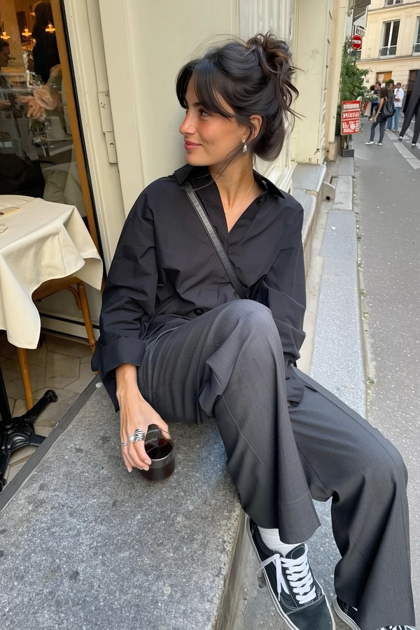 A candid iPhone-style photograph of a young woman naturally captured sitting relaxed at the edge of a bustling Parisian sidewalk café. She extends her legs comfortably along the textured stone pavement, holding a clear glass cup of black coffee in one hand. Her outfit features refined minimalist monochrome pieces from COS: a loose, soft black shirt and tailored high-waisted trousers with subtle creases showcasing fabric grain, paired with classic worn Vans sneakers. Her dark hair is styled effortlessly into a messy bun with delicate, tousled strands escaping naturally around her face. Bold yet understated silver rings adorn her fingers, adding a quiet statement that complements her casual look. The natural daylight casts gentle soft shadows and subtle highlights, authentically illuminating her skin texture, light freckles visible beneath minimal makeup, and the tactile details of her fabric and accessories. The scene includes typical Parisian sidewalk café elements: worn stone pavement, a small round table partially cropped with a muted neutral tablecloth, and subtle street life softly blurred in the background. The framing is informal, slightly tilted and off-center, reflecting the spontaneous, intimate aesthetic of candid iPhone street photography.