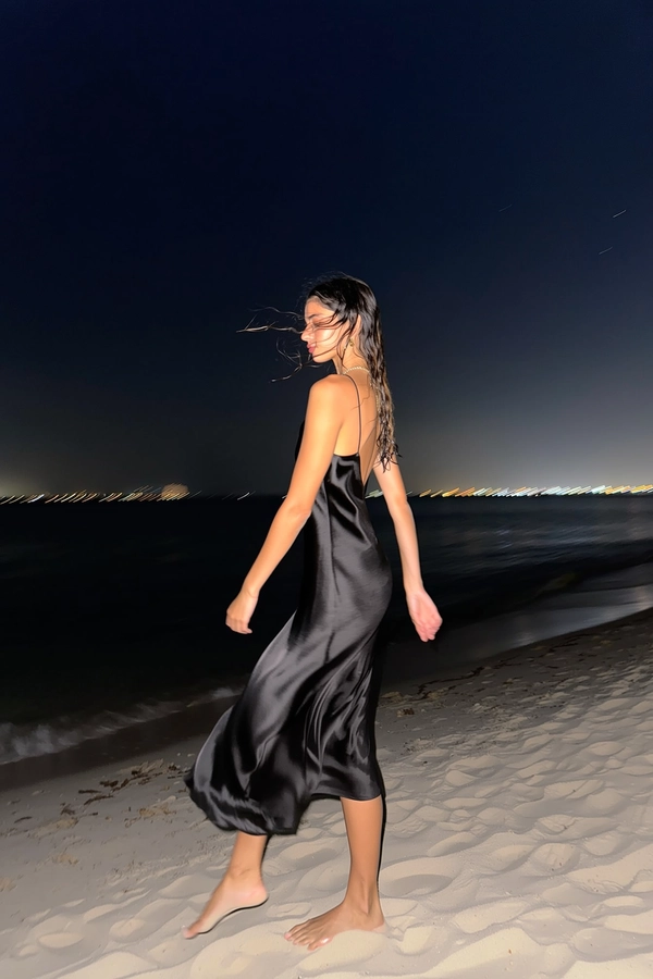 A sudden burst of iPhone flash pierces the enveloping night, revealing her tall silhouette clad in a flowing black satin slip dress that ripples gently with the seaside breeze. Her face, softly turned away, catches faint glimpses of distant city lights blurred far beyond the shoreline. Bare feet nestle delicately in the textured sand, as strands of damp hair drift and catch a subtle sheen under the sharp flash, contrasting the deep velvety darkness filling much of the frame. The slight blur at the hem merges with gentle wavelets, while expansive negative space embraces her poised calm, evoking silent sophistication and natural grace—flash-lit elegant beach snapshot, captured on iPhone.