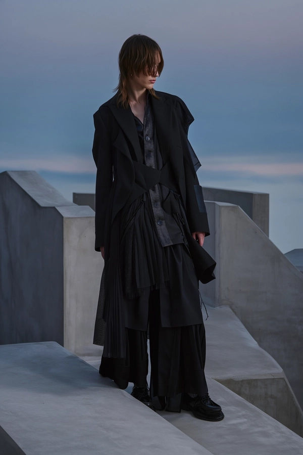 A quietly moody, dusk-inspired iPhone snapshot depicting a model dressed in an avant-garde outfit inspired by Junya Watanabe Comme des Garçons menswear. The ensemble features layered black fabrics with mixed textures, combining mesh and shiny materials to create a voluminous, architectural silhouette. Positioned slightly off-center near angular Brutalist concrete structures, the setting bathes in soft, diffused twilight lighting with muted greys and cool dusky blues. The model's relaxed asymmetry and neutral expression, highlighted by matte fabric folds, subtly contrast with the textured concrete surfaces exhibiting gentle imperfections. This elevated, gently tilted downward framing fosters an effortlessly intimate and contemplative mood, surrounded by calm shadowy blacks and charcoals, harmonizing avant-garde fashion with the tranquil minimalist evening ambiance.