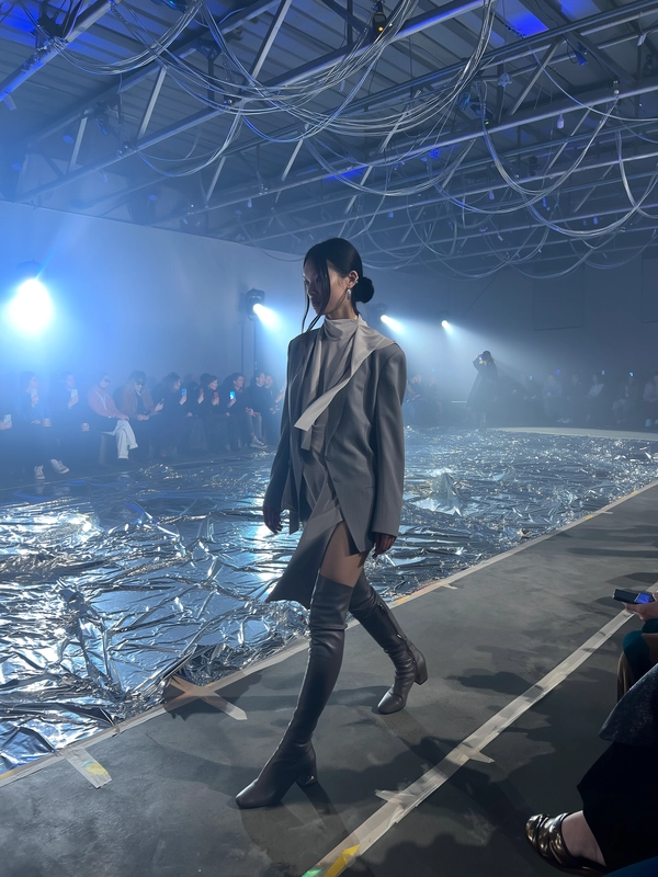 A model draped in layered muted greys and soft taupes crosses a surreal set strewn with crumpled silver metallic sheets catching reflections like moon craters. Her silhouette is relaxed yet precise, with oversized asymmetric tailoring brushing against thigh-high matte boots. Soft, cool blue backlights wash the industrial space into dreamlike hues, while sharp white spotlights carve subtle shadows over her calm, focused expression. The runway, a narrow strip of worn concrete, shows faint taped lines and slightly scuffed marks from heavy boots, blending tactile grit into the ethereal scene. Guests sit quietly along low benches, their blurred outlines softened by the dreamy haze as a few phones raise in the dim glow, blue light pooling against bare steel trusses overhead. The iPhone lens picks up gentle motion blur on the swinging metallic cords dangling from the ceiling, sensor noise in the deep shadows, and a delicate bloom around the soft LEDs that fill the space. Snap taken from an oblique front-row angle, with a slight hand-held shake and an intimate tight crop that draws you inside the lunar dreamscape—contemporary runway snapshot, shot on iPhone