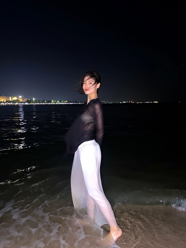 A delicate flash unfurls against the ink-black expanse, revealing her poised profile as she stands barefoot in shallow surf where city lights shimmer distantly across the quiet waves. Her sheer black top catches the faintest breeze, flowing softly alongside loose, fluid beach pants that ripple gently with the tide. Strands of dark hair fall tousled, kissed lightly by the night’s subtle dampness, while ambient reflections dance quietly on the water’s surface around her feet. The crisp sharpness of her calm expression anchors the scene, framed slightly off-center within velvety darkness that swells expansively, embracing an unhurried elegance and serene magnetism—flash-lit elegant beach snapshot, captured on iPhone.