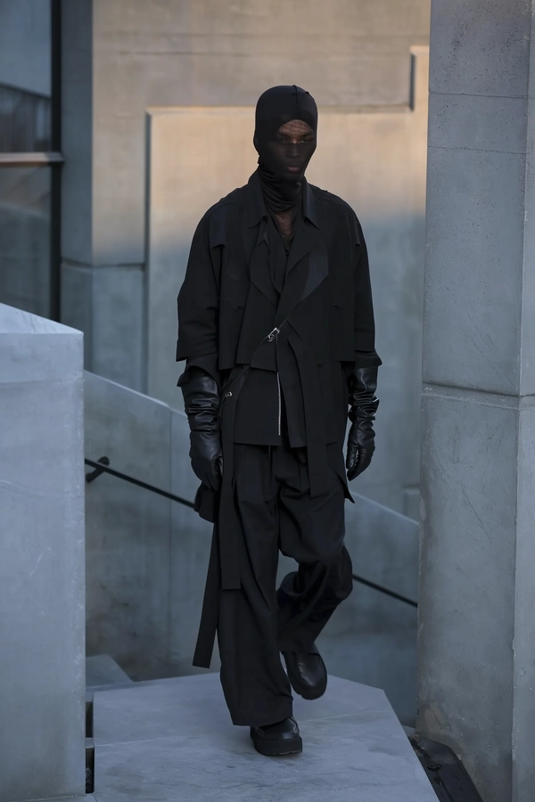 A softly dim-lit iPhone snapshot capturing a model walking slowly in an avant-garde Boris Bidjan Saberi ensemble composed of layered, predominantly black garments including a loose-fitting jacket, oversized pants, black leather gloves, and a semi-sheer head covering subtly obscuring the face. The model's expression is neutral, with minimalist hair and ultra-wide cheekbones hinted through the delicate sheer fabric. Set within a quiet Brutalist environment of gently weathered concrete walls in muted greys and steels-blues, angular geometric forms frame the background without direct sunlight. The softly diffused twilight casts subtle shadows and muted highlights, emphasizing matte textile folds and the quiet texture of the concrete surfaces. The composition is slightly elevated and tilted downwards, positioning the model casually off-center amid generous negative space, softly framed by textured concrete edges. The overall palette is calm and cool, replacing any crimson hues with shadowy black and dusky blues to maintain an effortlessly moody, intimate dusk ambiance.