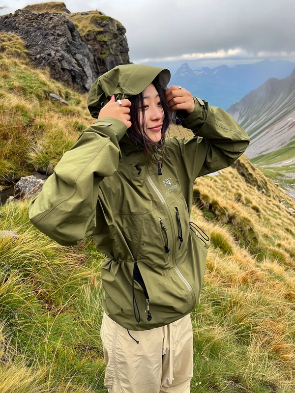 A candid, spontaneously captured iPhone photograph of a young woman standing relaxed amidst a rugged mountainous environment with grassy slopes damp from mist. She wears an olive green waterproof Arc'teryx hooded jacket with adjustable cords loosely tied, the durable technical fabric textured with natural creases and subtle wear, hood pulled up partially casting shadow over her softly relaxed face, damp strands of hair framing her cheeks. Beneath, she wears loose, beige pants that blend naturally with the environment. The scene features softly diffused daylight through partial clouds, gently illuminating wet grassy terrain and rugged stone outcrops. The photo emphasizes authentic textures: visible fabric water resistance, realistic grass textures with evening dew, and slightly weathered rock surfaces in the background. The framing is casual and slightly tilted, evoking the intimate spontaneity and rugged authenticity typical of stylish gorpcore outdoor iPhone photography.