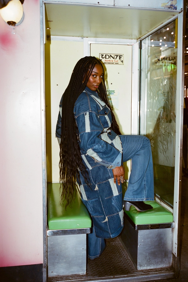 Set inside an intimate, timeworn photo booth adorned with faded avocado-green vinyl seats and mirrored chrome trims, a striking Caribbean woman commands the frame. Her long braids cascade with subtle sheen, darting shadows under the booth’s delicate sodium-vapour amber glow that spills softly on her skin, capturing the warm, rich undertones of her complexion. Clad in a meticulously crafted patchwork denim coat, each panel telling a story through varying shades of indigo and worn frays, paired with classic bell-bottoms that brush the textured rubber mat beneath her feet, she balances one foot up with fluid confidence. Her body twists slightly in a deliberate, poised stance, head thrown over the shoulder, eyes locked with the camera—fierce, unapologetic, breathtaking. The camera, positioned at eye-level with a 50mm lens, crisply frames the moment, the grain of the 35mm Kodak 5247 film stock lending an authentic tactile presence: from the slight pores on her skin to the rugged weave of denim fabric. The muted pastel palette, reminiscent of contemporaneous urban street snapshots, contrasts with the ambient reflections in the scratched glass panel beside her, composing a vivid tableau of retro realism and soulful grit. This is a nostalgic yet empowering homage to late-70s street style and individualism—late-70s / early-80s cinematic photograph, authentic film grain.