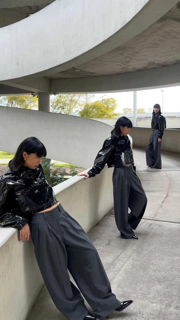 A surreal, candid iPhone-style photo capturing a woman duplicated multiple times along a brutalist spiral ramp inside an abandoned car park. She wears the same glossy cropped black jacket and ultra-wide charcoal trousers in every instance. Each figure is caught in a different casual yet natural pose — one climbing the ramp, another leaning lightly on the concrete railing, and a third mid-turn around a curve — interacting subtly with their surroundings like living architecture. Natural daylight filters softly through the open structure, casting realistic shadows and highlights that emphasize detailed fabric textures, hair strands, and skin imperfections. The framing is spontaneous and slightly unconventional, with unexpected crops and angles that amplify the surreal scale and depth of the repeating figures against the stark brutalist concrete, maintaining an authentic, unposed vibe characteristic of genuine iPhone snapshots.