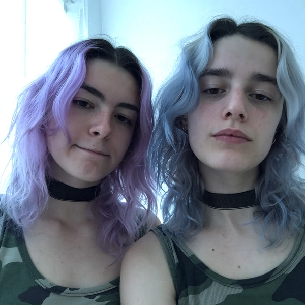 A dreamy, softly desaturated selfie of two pale young women with brightly dyed pastel hair—one with loose lavender waves and the other with tousled baby blue layers—both wearing distinctly mid-2010s chokers, one black mesh and the other paired with a faded camo tank top. Captured in natural, muted daylight filtering through a softly glowing window, the image showcases gentle cool-toned shadows in soft bluish-gray hues. The girls display authentic relaxed expressions and overplucked brows, with subtle minimal makeup and visible skin texture imperfections. Their casual composition includes a creatively informal centered framing with a slight tilt, evoking a candid snapshot style that resembles iconic Tumblr-era images commonly re-shared in 2015. Textures feature finely defined hair strands, cotton fabric grain, and choker material details, all unified with intentionally softened contrasts and muted color saturation, epitomizing mid-2010s spontaneous, intimate photographic realism.