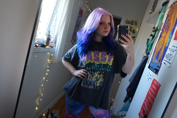 A candid mirror selfie of a young woman with vividly dyed pastel purple and deep blue hair styled in loose waves, wearing galaxy print leggings and an oversized graphic tee reading “Sassy Since Birth.” She stands casually in her cluttered hallway, one hand confidently on her hip, the other holding a cracked phone, surrounded by an eclectic backdrop of colorful posters, twinkling fairy lights, and handwritten song lyrics taped to the walls. Natural ambient daylight filters softly through a nearby window, complemented by subtle direct flash, producing intentionally muted color saturation, softly lowered contrast, and distinctly cool-toned shadows with soft lavender-gray hues. Detailed textures are apparent—the realistic grain of her cotton tee and leggings, delicate strands of her dyed hair, subtle freckles and skin imperfections, and the worn texture of her cracked phone screen. The composition is informally off-center with a slight tilt, casually cropped as if snapped spontaneously, authentically replicating mid-2010s candid smartphone photographs with a relaxed, intimate vibe.