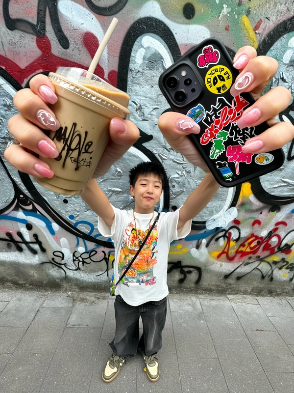 A casually chic, spontaneously captured iPhone-style image featuring a tiny, fashionably dressed young man wearing a bright graphic t-shirt and stylish sneakers, standing before a colorful urban graffiti wall. He extends dramatically elongated giant hands prominently toward the camera, holding an iced coffee cup and a smartphone adorned with bold, vibrant stickers. His relaxed, confident expression and modern short hairstyle complement his casual streetwear look. Soft natural outdoor light authentically illuminates detailed textures like fabric weave, glossy nail art, subtle skin imperfections, and sticker finishes. The slightly tilted and creatively wide-angled framing enhances the playful exaggeration of his oversized hands, reinforcing a contemporary, effortlessly stylish urban vibe typical of spontaneous lifestyle iPhone photography.