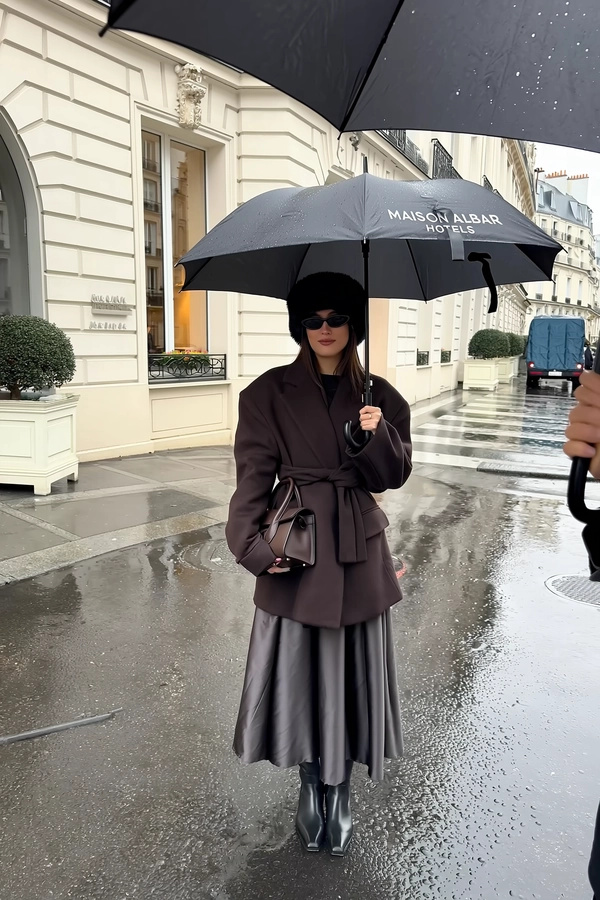 She stands poised beneath a sleek black umbrella branded "MAISON ALBAR HOTELS," its white lettering crisp against deep drizzle, dark brown oversized coat cinched neatly over flowing charcoal satin skirt; narrow black sunglasses shield calm eyes beneath a plush faux-fur hat, a small structured handbag hugged close under the canopy. The glossy Parisian pavement mirrors cream stone boutique hotel walls trimmed with tall windows and sculpted shrubs in square planters, wet surfacing catching soft daylight filtered through low clouds. Rain beads glisten on broad coat lapels and umbrella ribs, droplets tracing slight smudges on the lens as muted reflections ripple softly around her pointed black leather boots. The eye-level framing holds her central and still, umbrella edge casually cropping the top frame, handheld shake adding delicate motion, a quiet moment captured in a slick city rainscape—rainy city snap, shot on iPhone