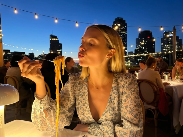 Under the dimming sapphire sky, a blonde woman in a softly patterned floral dress sits poised at a rooftop table adorned with glowing string lights. Her sleek, straight hair is tucked subtly behind one ear, revealing a calm countenance that veers neither smile nor frown, only complete stillness. The delicate fabric of her dress rustles gently with the evening breeze, lending an effortless elegance that harmonizes with the cityscape spread like a shimmering tapestry below.

She delicately lifts a fork, twirling strands of truffle-scented pasta glistening with earth-toned sauce, drawing it close to her slightly parted lips. A barely noticeable gloss catches the last warm rays filtering through the sky, highlighting the soft curvature of her mouth engaged in the quiet act. The deep umami sheen of the sauce glides across the richly textured noodles, inviting a subtle sensory pause caught perfectly in the frame.

Surrounding her, warm amber bulbs engineer a cozy, intimate ambiance as tables bustle quietly with patrons blending effortlessly into the fading light. The sharp silhouettes of urban towers punctuate the backdrop, their windows sparkling like distant stars, all framed by the delicate weave of string lights that bow gently overhead.

Captured from a gentle side angle as if by an arm’s length, the image holds a serene charm, every pore, wrinkle in fabric, and glint on glossy strands emphasized with natural clarity. This moment, perched delicately between casual and editorial, perfectly resembling a candid iPhone photo.