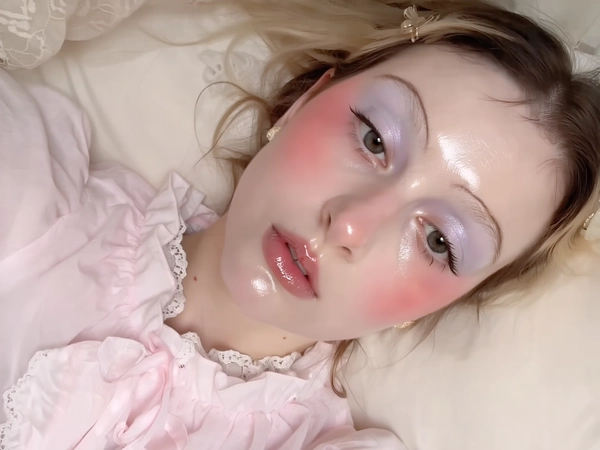 A close-up iPhone-style portrait of a young woman with milky fair skin displaying a natural, dewy glow with visible pores and soft skin texture. Her makeup reflects the babydoll style with heavy, exaggerated pink blush sweeping across her cheeks and beneath her eyes, blended seamlessly for a youthful flush. Her eyelids are covered in pearlescent pastel eyeshadow in soft shades of pearl white and pale lavender, catching the light delicately without harshness. Her lips are softly glossy in a gentle pink tone, capturing natural moist texture. Her eyebrows are natural dark brown, softly brushed upwards and shaped intentionally yet casually. The lighting is natural and soft, typical of iPhone photography with gentle window daylight diffused evenly across her face, avoiding hard, artificial shadows. The photo is casually framed from just above her head to her upper chest, slightly off-center and tilted to imply spontaneity. She wears a soft baby pink puff-sleeve blouse with lace detailing at the collar and delicate ruffle trims, the fabric showing subtle translucence and fine stitching. Her expression is relaxed and natural, lips parted slightly, eyes gazing gently off-camera. The background is a softly blurred pastel cream, suggesting a softly lit indoor environment with subtle textures like bedding or curtains. Fine hair strands and flyaways, natural skin pores, and glossy lip detail are clearly visible to emphasize authentic texture and tactile realism.