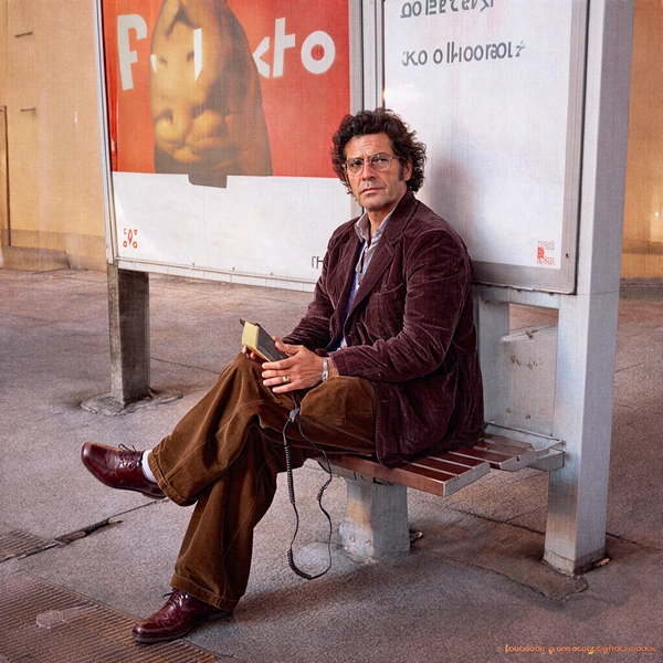 In the frame, a Mediterranean man commands attention with a poised, self-contained demeanor. His deep maroon corduroy blazer, tactile and ribbed under the soft amber flicker of a nearby streetlamp, contrasts richly against the warm sepia of his wide-legged brown trousers—a classic nod to early 80s tailoring. His dark, wavy hair is tousled yet deliberate, casting subtle shadows across his slightly sun-weathered face. With a cool yet distant gaze, his eyes wander beyond the immediate, reflecting a reflective solitude under the faded glow of an old advertisement billboard overhead—its peeling paper textures telling stories of a time-worn streetscape.

The setting evokes a late dusk hour bathed in sodium-vapour light, which bathes the scene in an amber haze that softens the bus stop's metal bench and the worn concrete underfoot. The Walkman he holds—a boxy, white Sony model iconic of the era—catches a gentle highlight, its crinkled leather strap adding an intrepid sense of tactile realism. His pose is relaxed but deliberate, sitting sideways on the bench with one leg crossed over the other, fingers loosely wrapped around the device, poised between motion and rest.

Filmed through a 50 mm lens at eye-level to capture intimate detail, the shot bears the grainy, tactile signature of 35 mm film stock, with visible gate weave that deepens the texture of his skin pores and the corduroy’s plush ridges. The composition balances the subject against the geometric austerity of the billboard frame behind him, the juxtaposition of human warmth against the cold, industrial urban environment.

Rendered in a palette reminiscent of Kodak 5247, the scene radiates a nostalgic golden-hour glow that encapsulates quiet urban solitude infused with 1980s street realism. The evocative lighting, coupled with subtle vignetting, enhances the mood of introspective cool. This carefully composed portrait channels the spirit of early 80s cinematic photography with authentic film grain, capturing a moment frozen in both time and emotion.

—late-70s / early-80s cinematic photograph, authentic film grain.