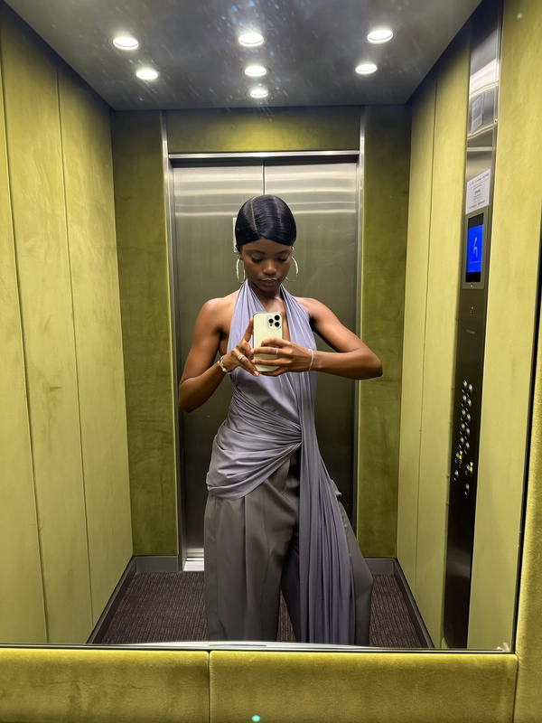 A delicate spread of moss green suede wraps the elevator cabin, its calming embrace softening the glow of the warm overhead lights. She stands centered, a long halter-neck dress in whispered chiffon layered over draping wide suit trousers that sway with an effortless rhythm. The phone is cradled gently at chest height, her fingers arranged with the quiet precision of a gallery catalog, as if each movement is a curated exhibition of relaxed refinement. A pale sheen catches the folds of fabric; her skin texture hums under the soft light, and a faint fingerprint smudge lingers just below the lens. The frame cuts slightly above the waist, as the elevator hums softly—a private moment of poised ease between floors as the closing doors hint at movement beyond—elevator mirror capture, urban chic on iPhone