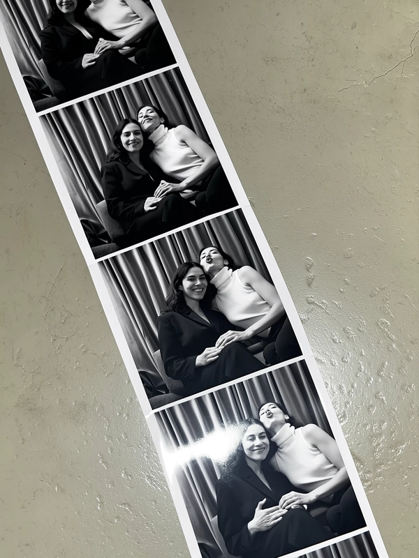 A vertical black-and-white photostrip curves diagonally across a matte, slightly scratched concrete surface, its subtle texture contrasting softly against the monochrome tones. Inside the booth, two women reside against heavy velvet curtains in deep muted gray, their backdrop textured and rich. The first frame shows them seated upright, poised yet casual, one in a wide-shouldered blazer dress, the other in a sleeveless ribbed turtleneck. Next, they lean affectionately into one another, sharing warmth and relaxed intimacy, hands gently resting or lightly touching. The final shot captures playful exaggeration: both offering dramatic, sultry pouts with slight head tilts. The strip carries the signatures of authentic photobooth prints—visible fingerprint smudges, faint creases softly bending the paper, and a delicate graininess overlaying each frame. Gentle iPhone flash reflections flicker across the surface, with shadows molding softly around their silhouettes. The whole composition tilts naturally as if casually grasped, evoking a candid, spontaneous vintage charm—contemporary photobooth snapshot, shot on iPhone.