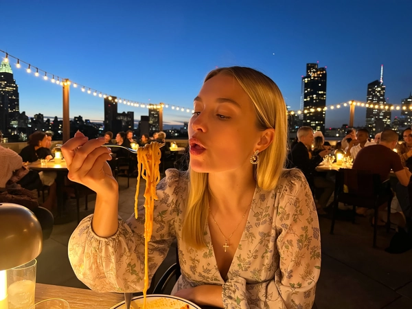 Under the dimming sapphire sky, a blonde woman in a softly patterned floral dress sits poised at a rooftop table adorned with glowing string lights. Her sleek, straight hair is tucked subtly behind one ear, revealing a calm countenance that veers neither smile nor frown, only complete stillness. The delicate fabric of her dress rustles gently with the evening breeze, lending an effortless elegance that harmonizes with the cityscape spread like a shimmering tapestry below.

She delicately lifts a fork, twirling strands of truffle-scented pasta glistening with earth-toned sauce, drawing it close to her slightly parted lips. A barely noticeable gloss catches the last warm rays filtering through the sky, highlighting the soft curvature of her mouth engaged in the quiet act. The deep umami sheen of the sauce glides across the richly textured noodles, inviting a subtle sensory pause caught perfectly in the frame.

Surrounding her, warm amber bulbs engineer a cozy, intimate ambiance as tables bustle quietly with patrons blending effortlessly into the fading light. The sharp silhouettes of urban towers punctuate the backdrop, their windows sparkling like distant stars, all framed by the delicate weave of string lights that bow gently overhead.

Captured from a gentle side angle as if by an arm’s length, the image holds a serene charm, every pore, wrinkle in fabric, and glint on glossy strands emphasized with natural clarity. This moment, perched delicately between casual and editorial, perfectly resembling a candid iPhone photo.