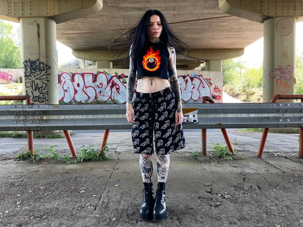 A heavily tattooed young woman stands confidently under an industrial overpass, framed by rough concrete pillars and rusty metal railings, captured in a full-body shot from head to toe. Her long black hair flows loosely, slightly tousled. She wears a cropped black sleeveless top featuring a bold, flaming 8-ball graphic, paired with loose black drawstring shorts adorned with a sharp punk-inspired logo print. Her pale skin is textured and natural, tattoos of skulls, roses, and gothic designs vividly covering her arms and legs with subtle imperfections visible. Heavy black platform combat boots ground her stance. Soft natural light filters through the overpass, casting diffused shadows over the gritty concrete floor and graffiti-stained walls behind. The mood is raw and rebellious with a strong alternative vibe, captured with the intimate spontaneity of an iPhone snapshot.
