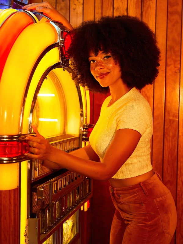 A warm, amber glow floods an intimate 1970s wood-paneled bar where a young mixed-race woman leans suavely against a chrome-trimmed jukebox glowing softly with translucent colored glass. Her voluminous natural curls form a halo around a freckled face touched by soft golden highlight, every pore and delicate fleck rendered with tactile precision. She wears a tightly hand-crocheted cream crop top whose fine nylon sheen contrasts starkly with the butter-soft rust suede bell bottoms, their rich nap catching subtle reflections of the sodium-vapour warmth that wraps the sagging honey-hued paneling behind her.

Her eyes lock boldly with the lens, sparkling with cool amusement and knowing confidence, framed by a slight smirk frozen in a moment of casual self-assurance. Captured at eye level on a 50 mm lens, the composition crisply delineates her expressive features while the smoky shadows of the bar blur discreetly behind. The film’s subtle gate-weave grain breathes authenticity into this 35 mm Kodak Gold stock frame, amplifying every tactile detail from soft skin textures through the glowing fabric.

Diffuse amber light spills directionally, casting gentle half-shadows across the silky fabrics and wooden surfaces alike, enriching their depth and natural warmth. The framing tilts just barely off-center, inviting viewers into a spontaneous yet deliberately constructed scene that feels cinematic and nostalgic, a fleeting fragment of bohemian nightlife flirtatiously preserved in analog grain.

This portrait pulses with playful intimacy and timeless 70s glamour, where natural beauty and elegant personal style clasp hands beneath gentle reflections of chrome and wood—late-70s / early-80s cinematic photograph, authentic film grain.
