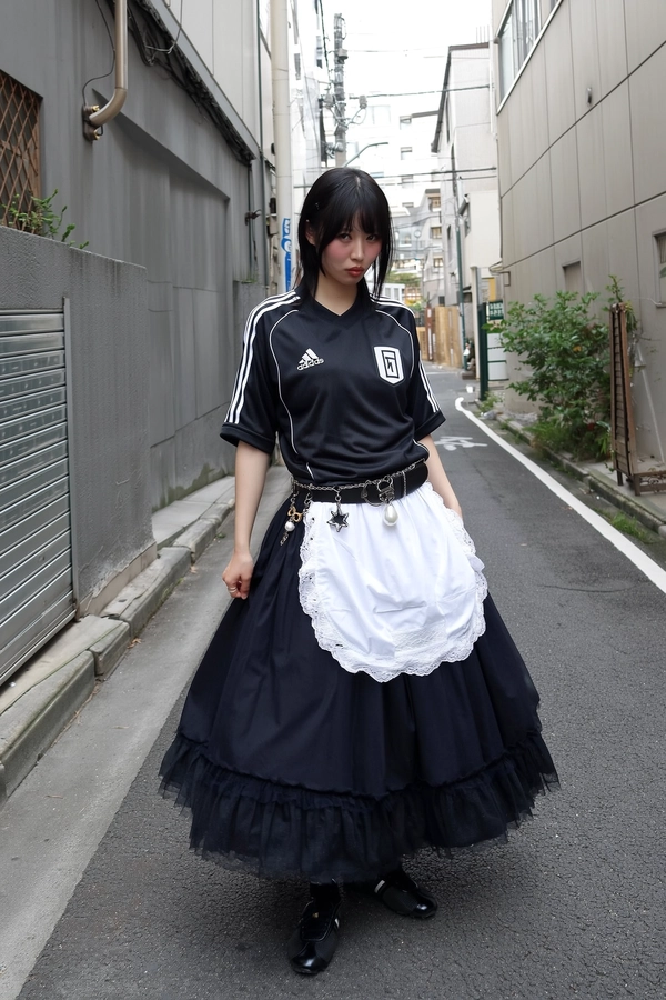 In a narrow Tokyo alley rimmed by cracked concrete and a raw metal shutter, a young woman stands poised in an oversized black Adidas football jersey tucked neatly into a flowing voluminous skirt. Draped over this, a stark white lace apron unfurls—its delicate fabric bright against the muted urban textures, hinting at playful defiance. A thick belt adorned with metal charms—a dangling star and pearl—circles her waist like a ritual relic, adding weight and personality. Her sleek black bob softly frames cheeks flushed with gentle blush, lips rest in a neutral pout, radiating quiet rebellion. Overcast daylight softens shadows and textures, lending mild sensor grain and muted tones to the scene. The iPhone’s handheld perspective catches her from a relaxed hip height with slight horizon tilt, infusing genuine spontaneity while the empty alley breathes urban isolation. Tactile details pop: matte fabric folds, delicate lace weave, charm metalinks, and rough pavement grit. An unexpected fusion of streetwear with gothic-lolita spirit, silently echoing Tokyo’s underground pulse—Tokyo street snap, shot on iPhone