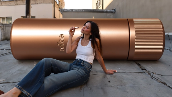 A spontaneous, candid iPhone-style photo capturing a young woman casually lounging back on a textured cement rooftop in an urban setting. She wears a simple white tank top with a subtle ribbed texture, paired with high-waisted flared jeans featuring visible denim weave and slight fading near the knees and hems. Her natural hair falls loosely around her shoulders with soft stray strands catching the gentle daylight. She leans comfortably against an enormous, hyper-realistic bronzer stick container—its sleek metallic bronze surface reflecting soft highlights and subtle wear marks, with detailed branding and finely textured cap edges. In her hand, she holds a smaller standard bronzer stick, which she applies delicately to her cheek, creating an intimate moment. Warm natural daylight softly glows from the side, casting gentle shadows across her skin texture, fabric folds, and the smooth cement rooftop sprinkled with faint cracks and scattered debris. The composition is slightly off-center, with informal framing cutting part of the bronzer container slightly at the edge, evocative of a spontaneous iPhone snapshot that melds everyday casual style with surreal scale exaggeration in a believable urban environment.