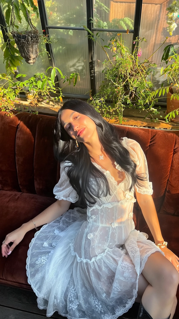 A softly intimate moment delicately revealed in warm, naturally diffused sunlight inside a sunroom lush with gently overgrown greenery, where a young woman reclines gracefully on a deep velvet chaise. She wears an ethereal Jenny Fax white sheer lace dress, intricately detailed with scalloped edges and delicate floral appliqués, featuring softly puffed sleeves and subtle ruffled layering. Her flowing black hair cascades loosely around her shoulders, complementing minimal accessories and fresh, natural makeup that highlights her serene, introspective expression. Golden shafts of afternoon light illuminate her face, enhancing the translucent lace textures and the rich velvet fabric beneath her. The gently weathered, plant-filled space adds quiet vintage charm, while the composition is framed from a subtle elevated angle with a slight tilt, creating a harmonious balance of tender femininity and effortless whimsical intimacy.