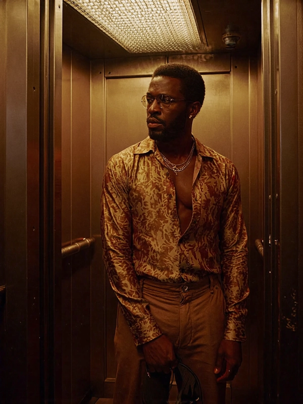 Bathed in the warm, moody amber glow of a retro office tower elevator, a Black man stands statuesque and solitary amid walls of glossy mirrored panels that slice the tight space into infinite reflections. His bold, sharply chiseled features catch the low, directional sodium-vapour light with sculptural clarity, while his intense, smoldering eyes cut through the glass, fixed on his own fiery image. His _silk_-patterned shirt hangs open at the chest—rich in sienna and burnt orange florals—that catches a subtle sheen underneath the vintage incandescent bulbs overhead, emphasizing the tactile luster of the fabric against his smooth skin.

Peer closer and the lenses’ shallow depth of field compresses the frame, pushing the mirrors into a dizzying geometric kaleidoscope around his powerful stance. The layered gold chains rest heavy and dull against the fluid folds of his shirt, contrasting crisply with the textured weave of his high-waisted brown flared trousers, reminiscent of ’77 Ralph Lauren tailored leisurewear. The elevator’s constrained space pulses with an analog grain reminiscent of classic Cinestill 800 Tungsten film, its soft gate-weave flickering faintly in the frame.

Shot on a 50 mm lens at eye level, the composition crops just above mid-thigh to capture his elegant, stoic posture—a stillness almost suffused with a silent narrative tension. This cinematic snapshot evokes a late-seventies aesthetic, reminiscent of Gordon Parks’ powerful humanist portraits, channeling an intimate yet electric mood of poised resilience, self-reflection, and stylistic bravado. The smudged fingerprints on the mirrored walls add a subtle tactile grit, grounding the surreal repetition in lived reality.

Overall, the frame radiates a nostalgic warmth through muted, earthy tones and carefully balanced highlights—the flicker of the elevator’s old wiring and the gentle hum of the city beyond implied in the scene’s silence. This is a timeless moment steeped in analog texture, metropolitan cool, and indelible presence.

—late-70s / early-80s cinematic photograph, authentic film grain.