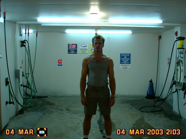 A sudden pop of on-camera flash blasts the man standing rigid inside the echoing car wash bay, framed dead center by dripping hoses and faded plastic signs. The tungsten-tinted fluorescent lights hum softly above, casting a cold, mixed glow that swirls in the hazy, slightly soft focus of the CCD sensor. Pixel fringing gnaws around the edges of his tucked-in tank top and tailored shorts, while JPEG blocks scramble the concrete floor beneath. His steady, straight-on gaze locks with the lens as the frame wobbles slightly with a Dutch tilt, like a quick grab and shoot. In the corner, a flickering battery icon blinks beside a timestamp: "04 MAR 2003  23:18". The photo quality is low-resolution and somewhat grainy.—early-2000s digicam snapshot, grainy low-res quality