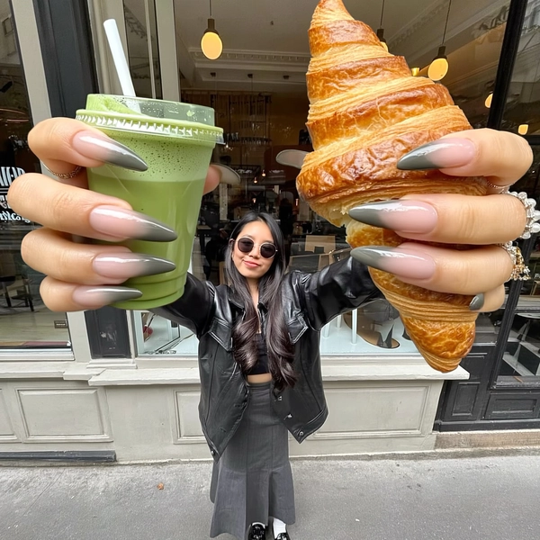 A casually chic, spontaneously captured iPhone-style image featuring a miniature young woman with long ombre nails and round sunglasses, dramatically extending oversized, elongated giant hands holding a matcha latte and a flaky croissant prominently toward the camera. She wears a sleek black leather jacket over a stylish skirt, exuding relaxed confidence and playful expression. The setting is a fashionable café exterior with subtle modern architectural details. Overexposed, natural film-style lighting softly illuminates the scene, revealing refined skin textures, genuine fabric grain, detailed nail art, and subtle jewelry finishes. The composition is casually tilted and creatively framed, emphasizing the exaggerated proportions of the giant hands while maintaining elegant balance and effortlessly chic authenticity typical of authentic iPhone lifestyle photography.