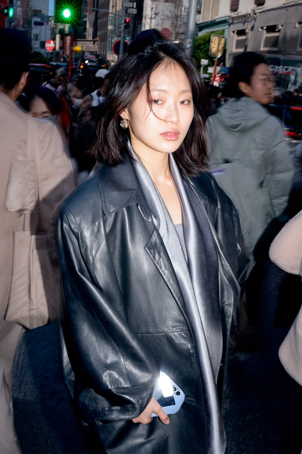 A Korean woman strides confidently through a dense Tokyo street, her sculptural leather jacket and soft-flowing scarf crisp against the chaotic hum. Harsh midday light slashes across her face, punctuated by direct flash that carves deep chiaroscuro shadows, highlighting the smooth grain of leather and the subtle pores along her jawline. Her phone is a quiet prop, sleek and luminescent in her hand, grounding the scene in modernity amid urban flux.

Blurred figures swarm around her in kinetic waves of motion, their forms melting into a pale bokeh that throws her figure sharply into relief. The scarf’s delicate weave and the jacket’s matte sheen absorb and shatter light differently, crafting a rich tactile tension between softness and edge. Her hair frames her face with a controlled tousle, while minimal, cool-toned makeup adds a polished restraint to the moment.

The backdrop is a gritty street scene distilled through a subtle tilt, compressing space as if time both slips and freezes. Shot from eye level on a 50mm prime at high shutter speed, the composition contrasts stillness and blur, framing her like an island of calm amid a sea of restless bodies. A faded Sakuran film emulation with slightly pushed contrast infuses the image with nostalgic grit and a modern pulse.

The palette drifts from cold urban steel to warm skin tones caught in a sharp flash flare, saturating details without losing intimacy—a visual poem of anonymity and urban coolness, distilled in fleeting focus and real skin texture.

—high-fashion editorial, hyper-real texture fidelity
