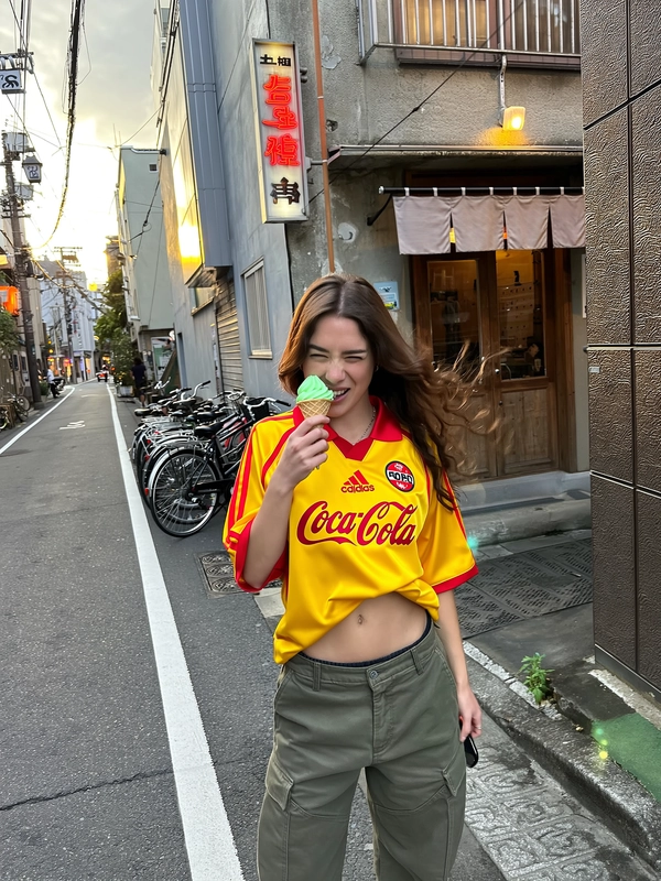In a quiet Tokyo backstreet where narrow sidewalks hug worn concrete walls and tangled overhead wires crisscross above, a young woman stands mid-bite of a vibrant green matcha ice cream cone. She rocks an oversized retro yellow Coca-Cola football jersey with striking red accents and a bold emblem, loosely tucked into low-slung olive cargo pants that reveal sturdy waistband details. Her long brown hair drifts softly with a natural flow as she smirks defiantly into the lens, embodying playful, carefree energy. Behind her, a classic sushi bar sports a weathered wooden door framed by a fabric noren curtain, while clusters of parked bicycles lean against textured walls. The soft evening sun casts a warm, cinematic glow that mingles effortlessly with subtle street shadows. The handheld, slightly tilted iPhone POV captures fractured light and spontaneous urban grit—worn pavements, cracked paint, and subtle lens flare from fading neon kanji, all converging in this candid moment where vintage sportswear meets Tokyo’s authentic street pulse. —Tokyo street snap, shot on iPhone