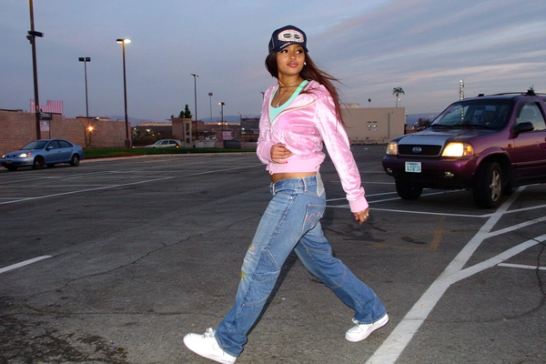 Mid-step crossing the parking lot, she rocks a glittery velour zip-up jacket flaring from the breeze, layered over a bright pastel tank top peeping out just right. Her low-rise, slightly baggy denim with subtle patchwork sways as she moves, the glow of car headlights stretching her silhouette long behind her. A rhinestone-studded Von Dutch trucker hat tilts back on her head, and chunky white Nike Air Forces hang on her feet, catching the cool evening light. The photo quality is low-resolution and somewhat grainy, the flash glimmer faintly reflecting off her rhinestones, capturing this spontaneous slice of early-2000s vibe with soft CCD camera grain and a casual tilt in framing. The whole scene feels genuine and free, just pure Y2K street style in motion.—casual candid early-2000s Y2K snapshot, grainy low-res softness