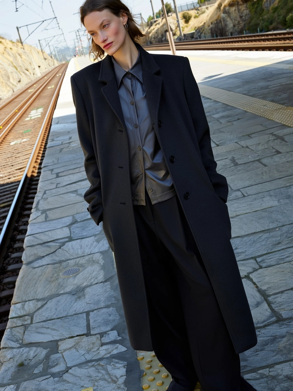 A spontaneous candid photo of a woman standing at a cliffside train station during soft daylight, highlighting her sharp cheekbones and porcelain skin with natural skin textures and light reflections. She wears a long charcoal overcoat with sculpted shoulders layered over a buttoned-up shirt and wide black wool pants, the fabric textures finely detailed. The rugged stone platform and minimalistic rail tracks offer an authentic natural setting. The image is shot with a casual, slightly tilted framing that conveys an unposed, intimate moment. The overall mood reflects the subtle ambient light and textural realism characteristic of authentic iPhone photography.