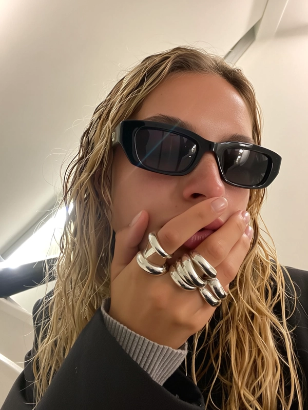 A young woman presses her wet-look blonde hair off her forehead, her hand stacked with bold, chunky silver rings—molten shapes twisting fluidly around her fingers—hovering close to her jaw. Glossy black square shades slant just enough to catch the faint reflection of an overcast sky, while the soft off-white backdrop highlights the fine pores and a faint lens flare rushing across the chrome edges. A ribbed cuff peeks from beneath a loose sleeve, fuzz catching the stairwell's faint fluorescent buzz. The subtle tilt of the frame and a tiny lens smudge make the intimate moment feel candid, capturing her minimal glam and aloof vibe. —close-up captured on Iphone, hand-face jewelry focus