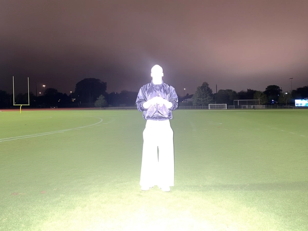 A lone Black man stands still in the center of a football field under a cloudy night sky, casually dressed in a glossy windbreaker and wide cargo pants. Captured with an intense direct flash from an iPhone, his figure is completely bleached out, facial features and clothing details obscured into a surreal, glowing white silhouette. The faint green of the field glows softly in the background, while the composition reflects the unplanned, candid feel of an amateur nighttime snapshot.