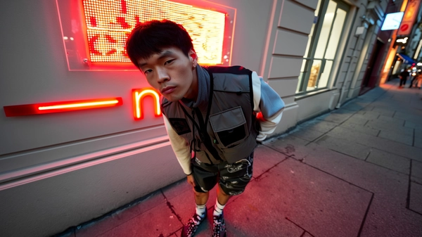 A dynamic, wide-angle iPhone-captured photograph features a young man leaning slightly forward with one hand casually in his pocket, his face bearing a tight, observant neutral expression. His sharply defined jawline is artfully highlighted by urban shadows, with lips gently sealed. He sports a creatively styled layered nylon vest featuring subtle tonal contrasts and functional detailing, paired with loose-fitting tech shorts designed with innovative textile textures distinct from any examples. His footwear includes uniquely mismatched socks adding a playful yet original touch. Behind him, the street scene glows softly under a vivid red LED sign spelling an abstract partial word “—end—”, casting gentle neon reflections onto nearby surfaces. The urban environment includes textured sidewalks and subtly illuminated building facades adding layered depth. Natural ambient evening light combined with the neon glow creates realistic, soft contrasts and shadowing. The image is framed with elevated camera angle and notable wide-angle distortion typical of spontaneous iPhone photography, emphasizing his engaged pose and direct eye contact with the lens, capturing authentic casual urban style and atmosphere.