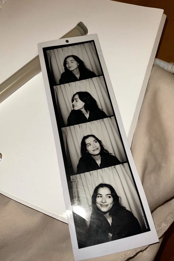 A narrow black-and-white photostrip rests casually atop a soft beige cotton throw, lying beside a simple notebook and pen, contributing to everyday warmth. The three frames capture one woman alone, her eyes closed peacefully in the first, glancing sideways with a hint of curiosity in the second, then settling into a relaxed, gentle smile in the third. Behind her, neutral curtains with faint folds create an unassuming photobooth backdrop. Subtle sensor grain and soft iPhone flash reflections lend quiet authenticity, while gentle creases and light fingerprint smudges reveal the strip’s tactile history. From a slightly skewed overhead angle, the composition feels candid and intimate—contemporary photobooth snapshot, shot on iPhone.