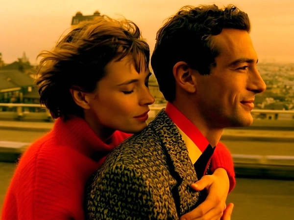 Amidst the gentle blur of a moving cityscape, a woman with short, tousled hair leans close to a man, her eyes tenderly closed as if savoring a quiet connection. Her vibrant red sweater glows warmly against the golden ambient light, which cascades over their skin in soft, honeyed tones, infusing the scene with nostalgic calmness. The man, wearing a patterned jacket with a bright collar, faces forward with a serene, slight smile, the strong contours of his profile catching the ambient glow. The close, intimate framing captures the whispered closeness between them, textures of fabric and skin rendered with a delicate film grain that evokes a timeless, romantic mood. The background's subdued motion contrasts with their stillness, enveloping the moment in a softly blurred embrace of movement and silence. —intimate contemporary cinema