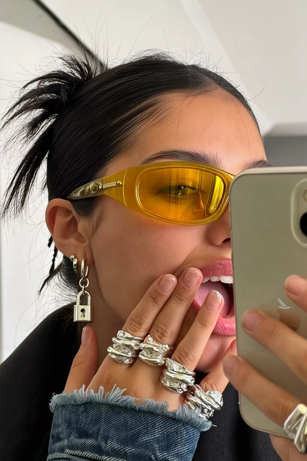 Golden strands rebelliously escape a messy bun as she presses her fingertips—stacked with chunky molten silver rings—near frosted lips, where subtle gloss barely catches the midday stairwell’s cold fluorescent glow. The yellow-tinted lenses curve around her face, reflecting an iPhone’s faint reflection in neat lock pendant’s shimmer. Tiny skin pores texture the sun-kissed cheek, contrasting with brushed metal ripples blurred softly behind her. Denim cuff fuzz peeks near the wrist, and the frame slants just enough to catch this unstudied moment, where molten silver forms dance in the ornate collage of a modern muse. —close-up captured on Iphone, hand-face jewelry focus