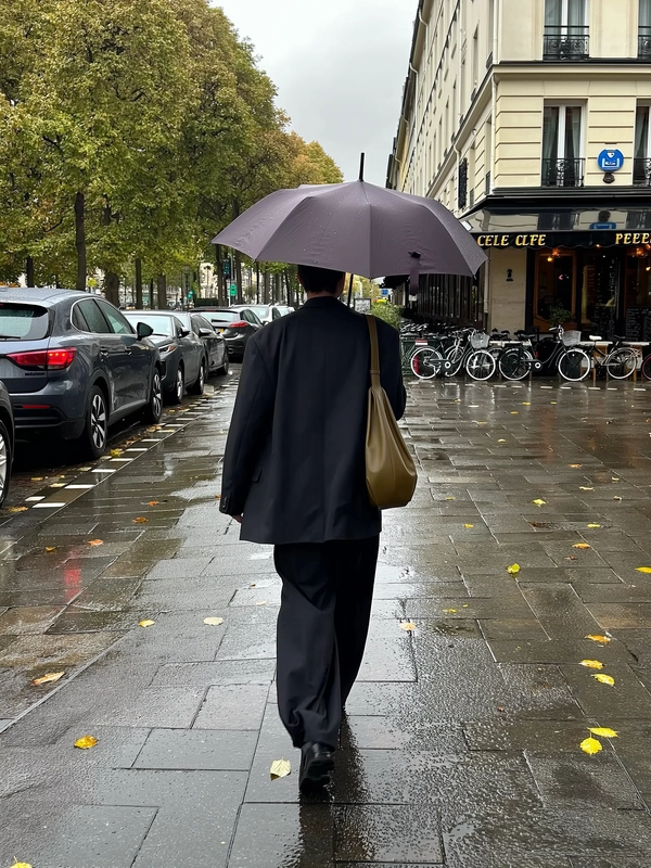 A single figure strides away under a large dark umbrella, oversized black blazer draping loosely over wide-leg trousers and dark shoes, a slouched olive-brown leather shoulder bag dangling at their side. The wet European sidewalk glistens with puddles and scattered yellow-green leaves, parked cars and bicycles resting under tall leafy trees to the left, while to the right a light façade classical building anchors the scene, its black café awning gilded in gold lettering. The cool, overcast daylight softens shadows and mutes colors into gentle neutrals, the pavement reflecting every delicate splash and ripple. The iPhone’s sensor gently captures rainbeads clinging to the umbrella’s ribs, a slight shake hints at a handheld vertical follow-shot from behind, centered on the solitary walker moving into the quiet distance—a calm, rainy city moment layered in minimalist style and reflective mood. —rainy city snap, shot on iPhone