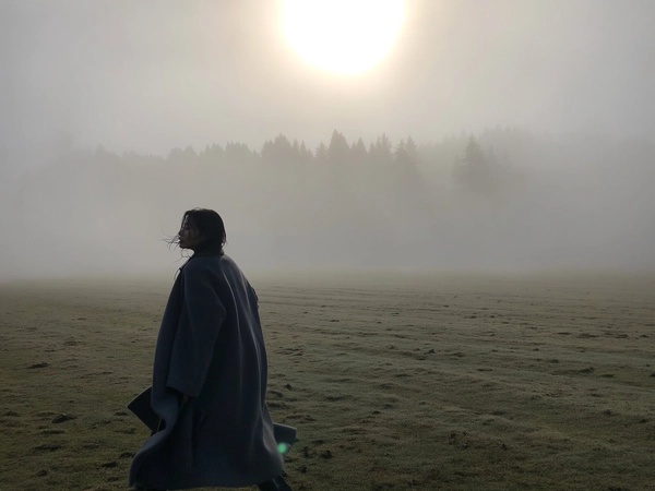 A solitary woman draped in a dark, oversized wool coat moves slowly across a dew-slick, fog-drenched field, her breath visible in the chill air as the mist curls around her silhouette. She turns her head back just slightly, loose strands of damp hair clinging to her cheek, while the blurred shapes of distant trees dissolve into the thick, pearl-grey haze. The soft light of an overcast dawn seeps through the low cloud, cooling the muted earth tones and casting gentle shadows that dissolve at the edges. The iPhone’s lens captures a tender grain in the muted shadows, a faint lens flare from the hidden sun, and a slight blur in the fluttering hem of her coat, all framed at waist-height with generous empty space ahead, amplifying the hush and introspection of the moment—mistbound reverie, shot on iPhone.