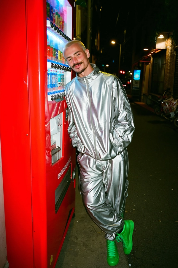 A candid, spontaneous shot of a man with a bleached buzzcut and sharp high cheekbones standing casually beside a glossy bright red vending machine in a dimly lit urban alley at night. He wears a metallic silver tracksuit featuring visible origami-style folds and bright green high-top sneakers, with the night ambient lighting casting soft reflections on the metal and fabric textures. The natural skin tone and pores on his face are visible, and his relaxed expression enhances the intimate, unposed feel. The photo is casually framed with a slight tilt, typical of an authentic iPhone capture highlighting genuine nighttime urban ambiance.