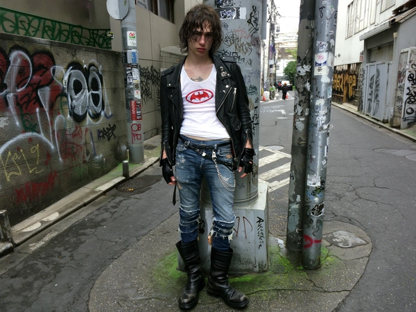 A slim young man stands against a graffitied utility pole on a cramped Tokyo backstreet, framed in a mid-close candid iPhone shot with a relaxed, off-center composition that crops just below his boots. His messy, damp curls fall unevenly around a pale face marked by subtle skin texture and slight redness. He wears a tight white tank top stretched slightly across his chest, emblazoned with a faded red bat logo, layered under an open black leather biker jacket with silver zippers and worn creases. His distressed skinny jeans, patched with fraying denim and held together by safety pins, sag low on his hips, a tangled metal chain swinging from a belt loop. Fingerless black gloves cover his hands, which display faint tattoos creeping across knuckles and wrists.

The background buzzes with raw urban energy: sticker plastered poles, peeling graffiti tags, and chipped concrete walls typical of gritty Tokyo alleys. His bulky black boots bear scuff marks, resting on cracked pavement dappled by soft, overcast daylight streaming through narrow spaces above. The light diffuses gently, casting muted shadows and a subtle film grain across textures that feel lived-in and immediate. The entire image pulses with early 2000s indie sleaze and emo punk street fashion, caught with genuine spontaneity, authentic imperfect framing, and a desaturated, natural color palette embodying underground urban Japan vibes.