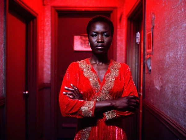 In a shadowy, intimate hallway of a 1970s jazz club, a statuesque Black woman stands poised and immovable, her arms firmly crossed beneath the soft glow of moody crimson lighting. She wears a sumptuous red velvet wrap dress, finely embroidered in shimmering gold thread, the textured fabric catching sparse light and revealing delicate folds that emphasize its sumptuous weight. Her skin, dark and radiant, absorbs and reflects the saturated rubescent hues with a velvety sheen that contrasts strikingly with the cool shadows creeping from the hallway’s worn paneling and faintly peeling wallpaper.

The atmosphere is thick with grainy 35 mm texture, giving a palpable analog authenticity as tiny gate weave flickers across the frame, while the light almost clings to the polished gloss of the ornate metallic embroidery that snakes along her neckline and wrists. Her expression is regal and unyielding, eyes steady and distanced—not quite inviting, yet commanding reverence. Her closely-cropped hair frames her face with precision, catching the sparse light that reveals subtle skin pores and the hint of matte makeup accentuating her natural features.

Shot from an eye-level 50 mm perspective, the composition centers her slightly off-axis, with the hallway receding diagonally behind her, deepening the shadow-play and drawing focus to her figure’s strong silhouette. The saturated reds bleed gently into near-black recesses, evoking the rich, tension-filled mood of early-80s urban nightlife scenes, reminiscent of cinematographers who thrived on chiaroscuro and tactile realism.

This evocative still channels the visual gravitas of early 1980s 35 mm cinema, where shadowy interiors and richly textured costume design meet the raw dignity of character study—an enduring image of strength and silent command under the soft burn of red sodium vapor-like glow.

—late-70s / early-80s cinematic photograph, authentic film grain.