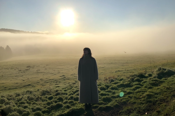 A lone woman cloaked in a high-collared, woolen coat stands motionless at the verge of a dew-laden meadow, her breath mingling with the thick, pearly fog rolling over distant hills barely traced behind her. The coat's textured fibers grip subtly damp air, strands of her hair loosening and damp against her neck as she gazes slightly away, absorbing the muted silence. Dawn's faint wash seeps through, casting gentle pastels and cooling green-browns into the soft blur that bleeds the horizon. The iPhone’s grain whispers in shaded corners, a faint flare skirts the pale sun veiled above, and the frame tilts just shy of eye-level, cradling generous mist-filled space around her—edges dissolving like a whispered secret. The quiet rustle of grass underfoot and a lone crow’s call brush the hush. Her solitary figure becomes a still poem of inner calm amid a softened world—mistbound reverie, shot on iPhone