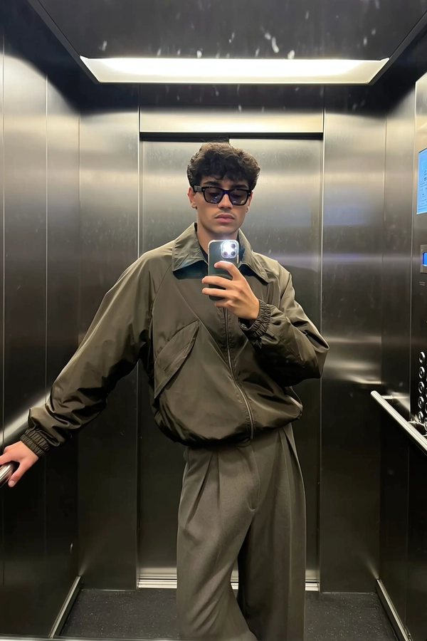 Matte olive panels soak in the shadow play, wrapping around him like a stage as he leans with arms stretched wide, fingers tracing the cool elevator rails. His collarless zip jacket, cut clean and crisp, catches a satin whisper under the overhead fluorescents, while wide-leg wool trousers fall with a soft gabardine weight that pulls the frame’s texture into quiet relief. Slightly cropped, the iPhone rests at chest-level, the screen glow illuminating a neutral, unreadable gaze locked on his reflection—not the lens. Fingerprint smudges ghost the glass, framing an unbothered stance staged between floors, the subtle crease of fabric and shadow folding together into a casually poised chiaroscuro. There’s a faint blur where the elevator doors begin to close, lending motion to the restrained luxury of effortless wayfarer style—elevator mirror capture, urban chic on iPhone