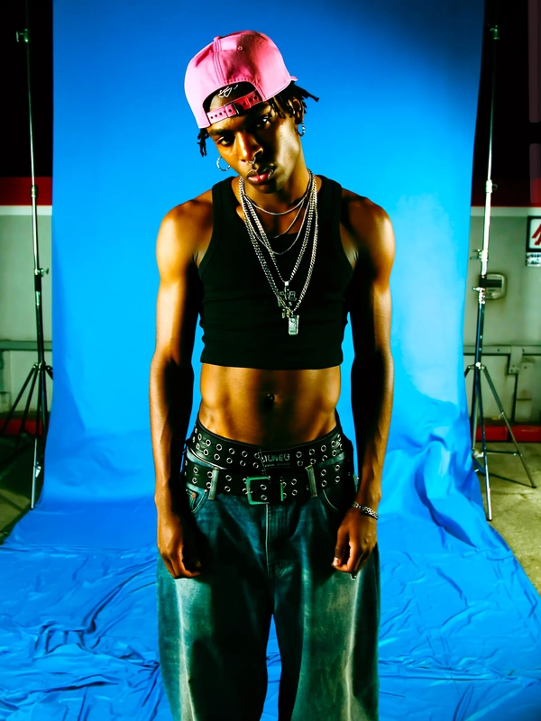 It’s a medium shot under the harsh fluorescent glow of a gas station at night, the cold light casting stark shadows across his face and cropped basketball tank top. His oversized jeans sag low, a thick black belt with silver grommets visible above the waistline, adding weight to the look. A fitted cap is turned backward, its bubblegum pink color clashing vibrantly against the black tank, while a pair of silver hoops glint on his ears. His skin tone reveals a biracial heritage, and his head tilts downward with his eyes peering upward at the camera, creating a brooding, almost confrontational mood. His lips are neutral, and layered chains hang loosely but deliberately around his neck. The background is a matte electric blue vinyl backdrop, slightly wrinkled on the floor, adding to the staged yet raw vibe. The image resonates with early Y2K streetwear aesthetics mixed with urban grit but captured in a studio-inspired setting that feels both authentic and stylized.
—casual candid early-2000s Y2K snapshot, grainy softness