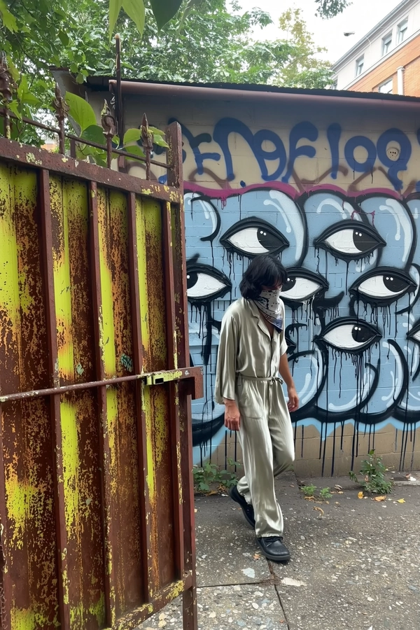 A candid iPhone-style street photograph capturing a tall figure walking past a rusted metal fence covered in peeling paint and rust spots. The person wears a loose, flowing silk jumpsuit and a painted scarf partially covering their face, creating a mysterious urban aura. Behind them, a graffiti-covered wall features several large, stylized eyes, one of which aligns strikingly with the figure's own gaze, establishing a surreal, intimate connection. Natural daylight filters softly over the scene, emphasizing the weathered textures of the rusted fence, chipped concrete, and layered graffiti paint drips. The framing is casual and slightly tilted at eye level, producing an authentic, spontaneous snapshot typical of candid street photography. The overall mood blends genuine urban rawness with a subtle sense of surreal street art interaction, rendered in vivid, realistic detail replicating the qualities of an iPhone camera capture.